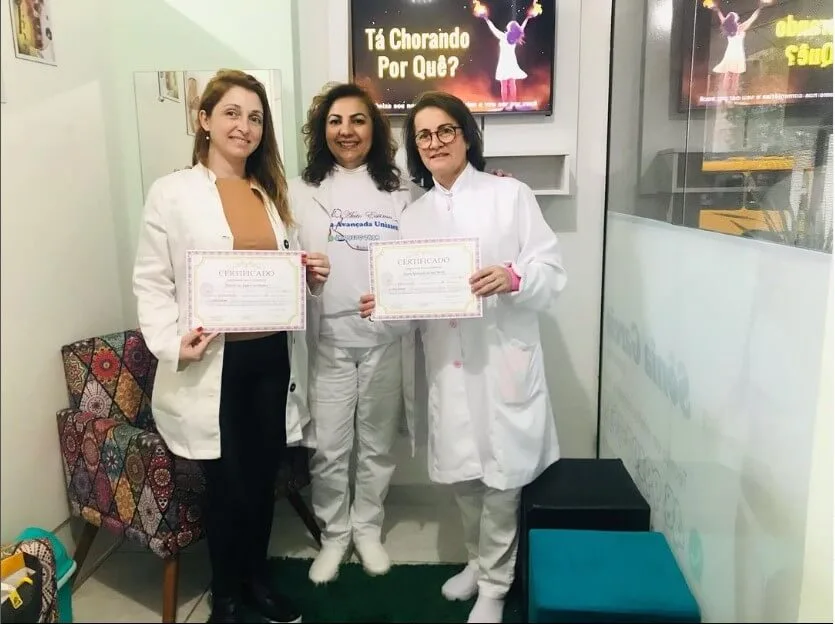 Três mulheres de jaleco branco, duas segurando certificados, na recepção da clínica